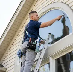 Professional window cleaner on a ladder using a squeegee to clean an exterior arched window, Cambridge, MA.